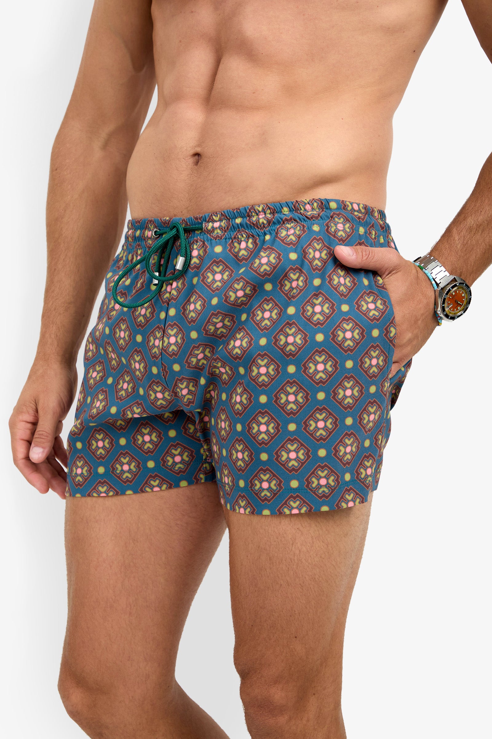 A man wearing the OCEANSIDE SWIM TRUNK, featuring a blue and multicolored geometric design, stands with one hand in his pocket and a watch on his wrist.