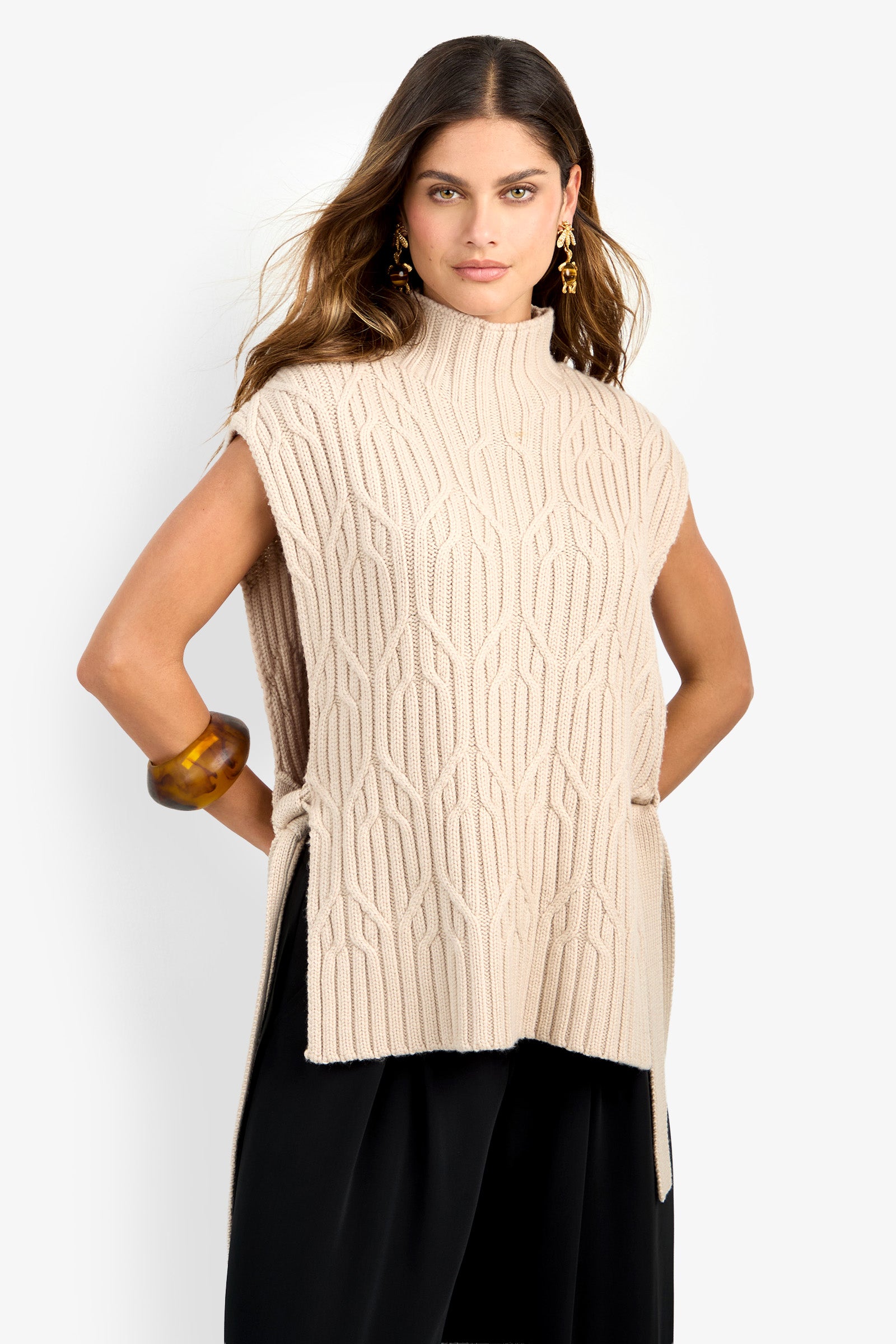 A woman stands against a plain white background, wearing the ROMEA CAPE—a sleeveless, beige textured knit sweater crafted from 100% wool with a high neck and side slits—paired with black pants, gold earrings, and a chunky brown bracelet.