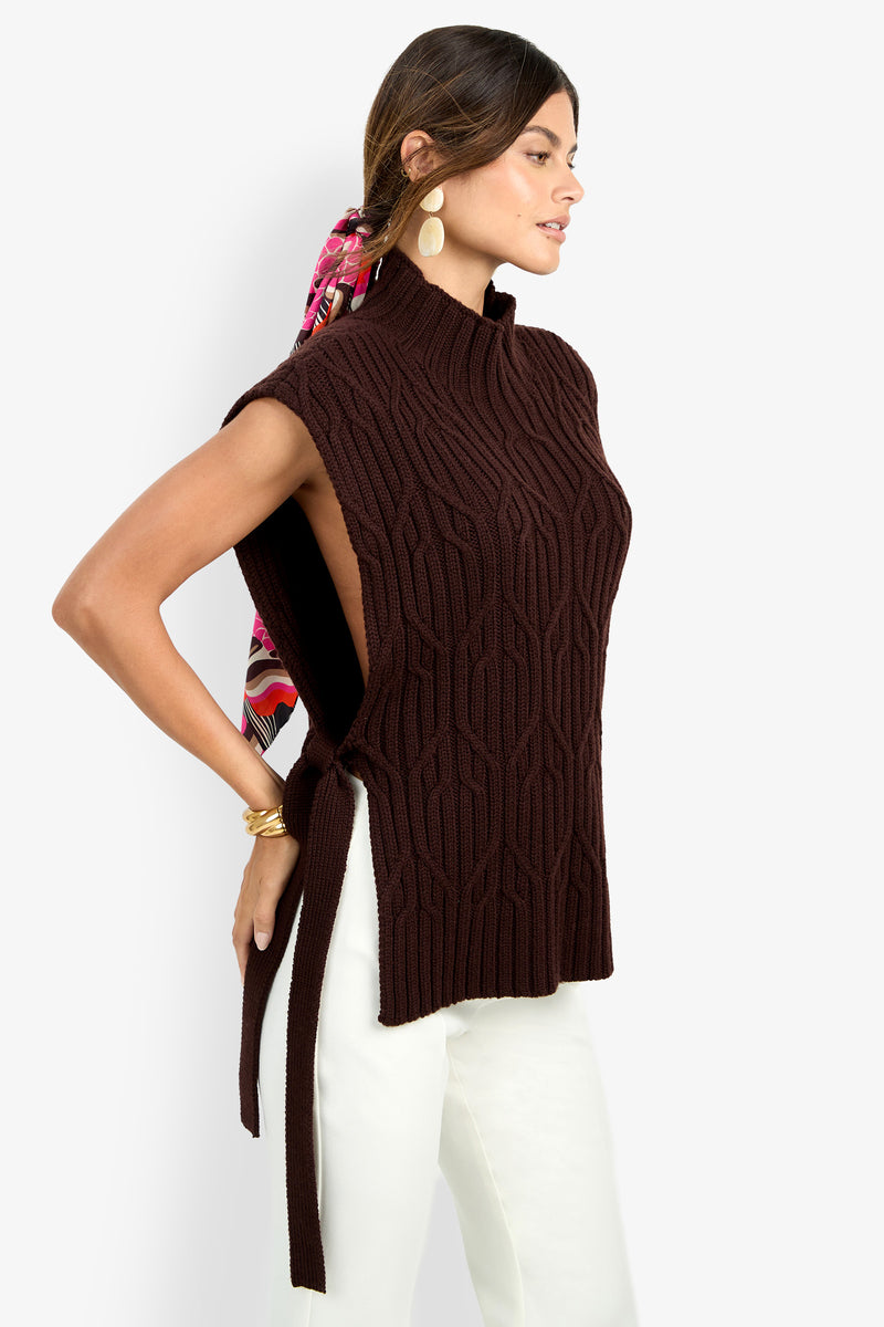 Woman wearing the ROMEA CAPE—a sleeveless, dark brown pure wool knit sweater vest with side ties—paired with white pants, large oval earrings, and a colorful scarf in her hair, posing sideways against a white background.