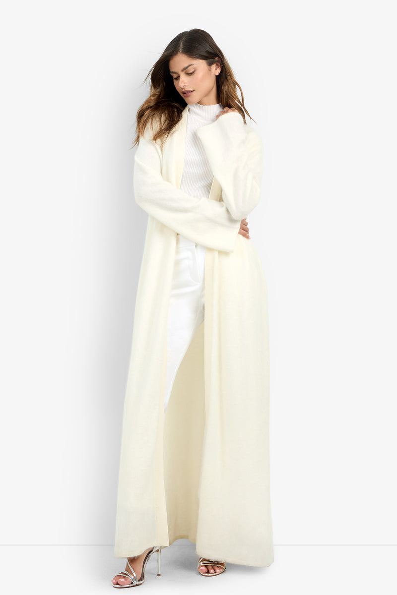 A person models a long, open-front almond Carducci Cardigan over a white mock-neck top and trousers.