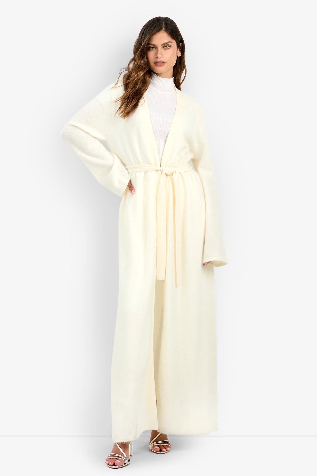 A person wearing a long, cream-colored, belted Carducci Cardigan over a white mock-neck top and strappy heels.