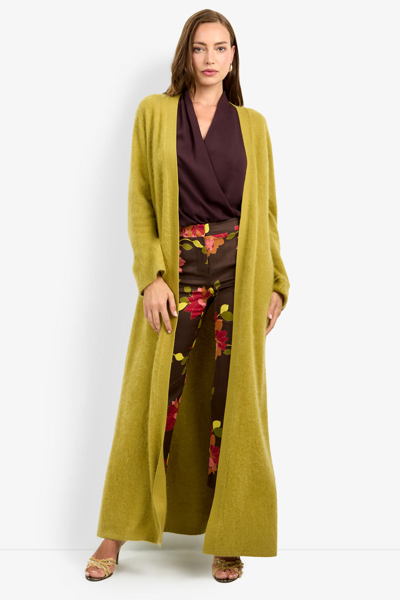 A woman wearing the CARDUCCI CARDIGAN in mustard-yellow, paired with a dark purple blouse and brown floral pants, stands facing forward with one hand resting on her cardigan.