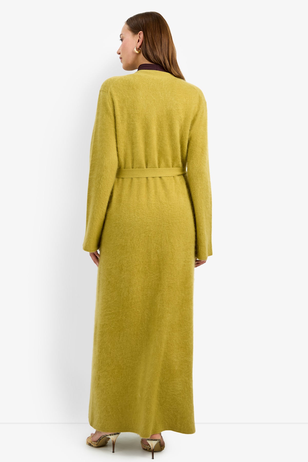 A woman faces away, wearing the CARDUCCI CARDIGAN—a long, yellow-green belted wool coat—paired with high-heeled shoes, and standing against a plain white background.