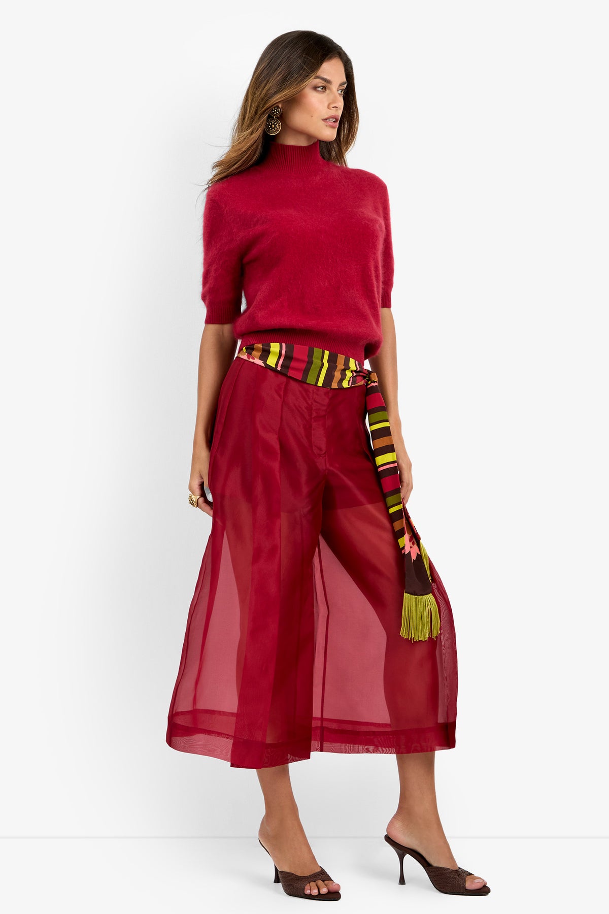 Woman wearing the Narni Short Sleeve Pullover, sheer wide-leg red pants with a colorful striped belt, and brown heeled sandals, standing against a white background.