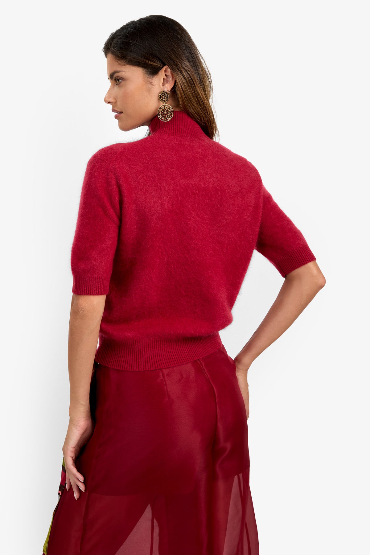 A woman with long brown hair, standing with one hand on her hip, faces away while wearing the Narni Short Sleeve Pullover—a red short-sleeve turtleneck sweater—paired with a sheer red skirt and large circular earrings. The background is white.