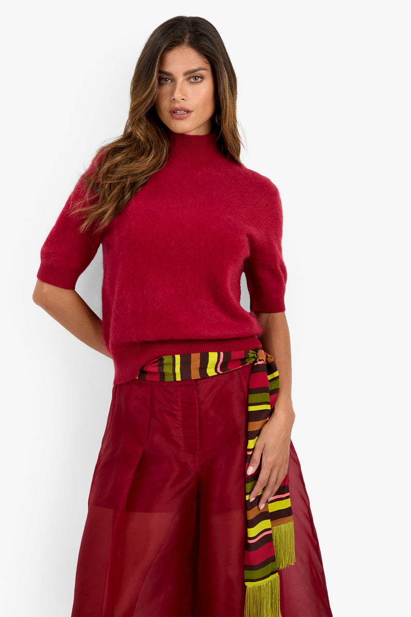 A woman stands against a plain white background wearing the Narni Short Sleeve Pullover in red, paired with a red sheer skirt and a striped, multicolored scarf tied at the waist.