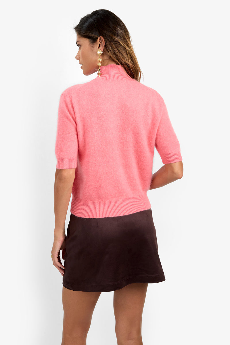 A woman with long hair, seen from behind, is wearing the Narni Short Sleeve Pullover in pink paired with a dark brown mini skirt and a dangling earring, set against a plain white background.