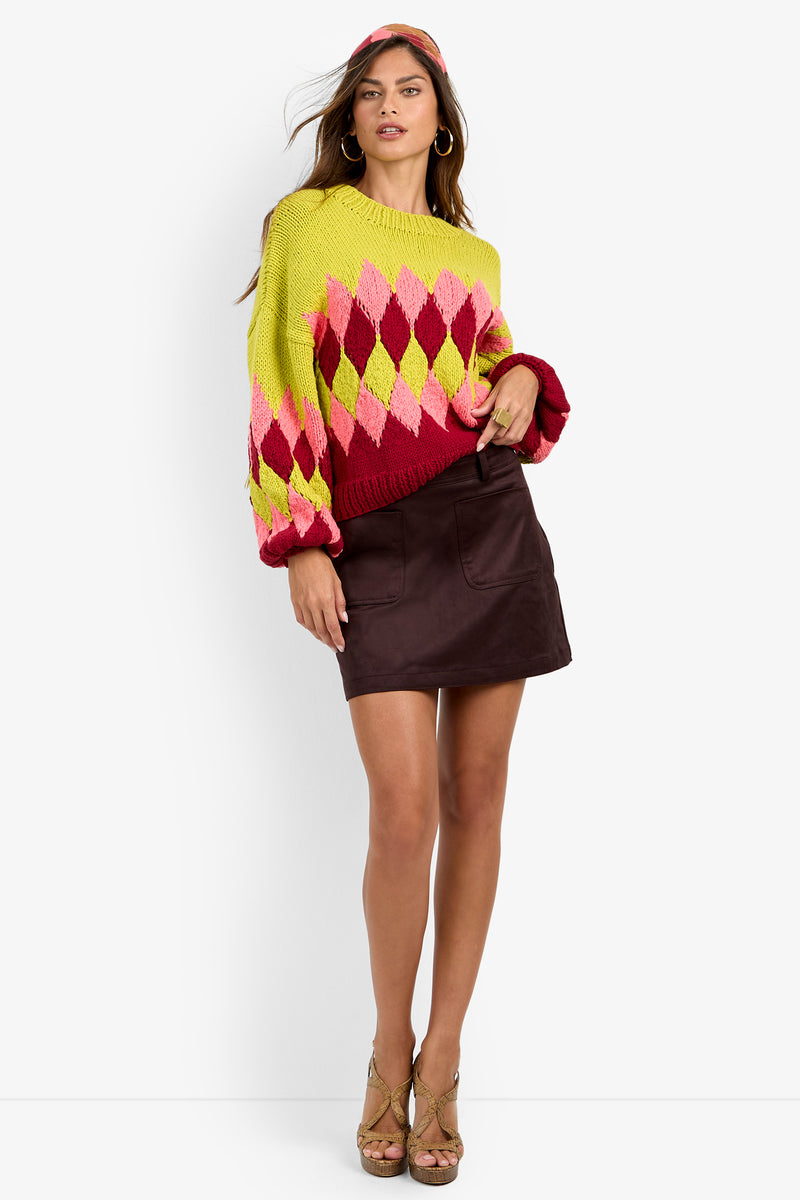 A woman stands against a plain white background wearing the Cavallari Sweater—an argyle knit in yellow, pink, and red—paired with a brown mini skirt, gold heels, and a matching headband.
