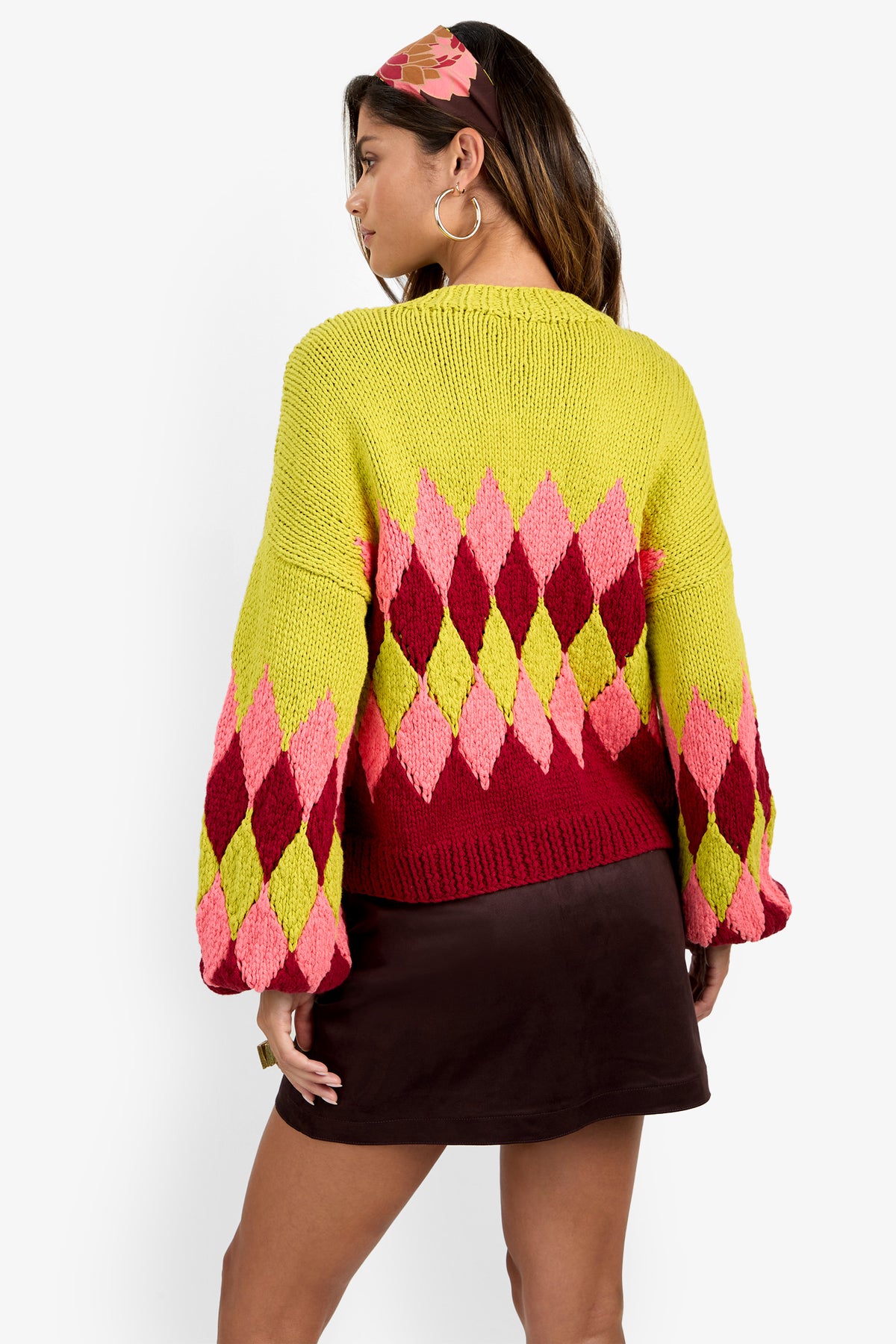 A woman with long hair wears the Cavallari Sweater—a colorful argyle knit in yellow, pink, and red—paired with a dark mini skirt. She is facing away to show off the back of the sweater, accessorized with large hoop earrings and a decorative hair accessory.