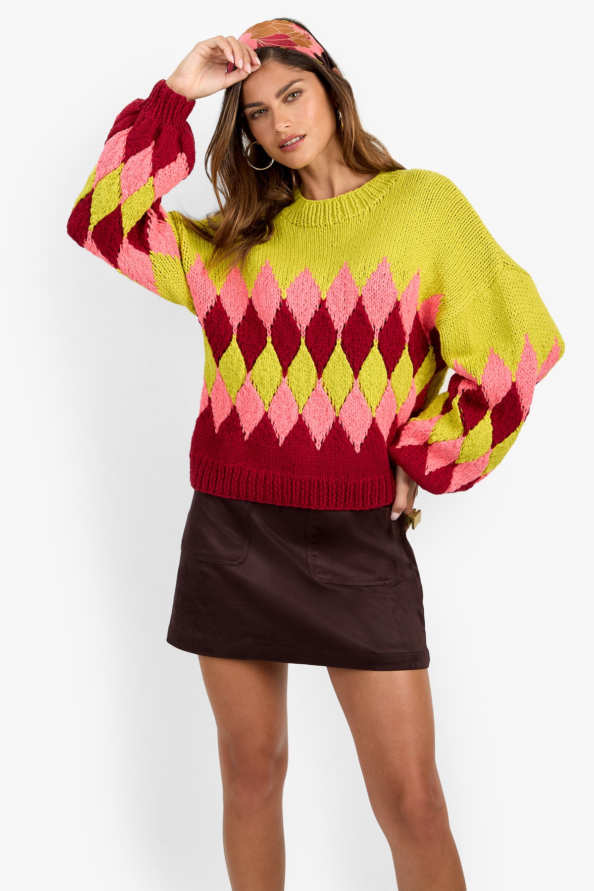 A woman is wearing the Cavallari Sweater—a colorful argyle knit in shades of pink, yellow, and red—styled with a dark brown mini skirt and a matching headband, as she poses with one hand adjusting her headband.