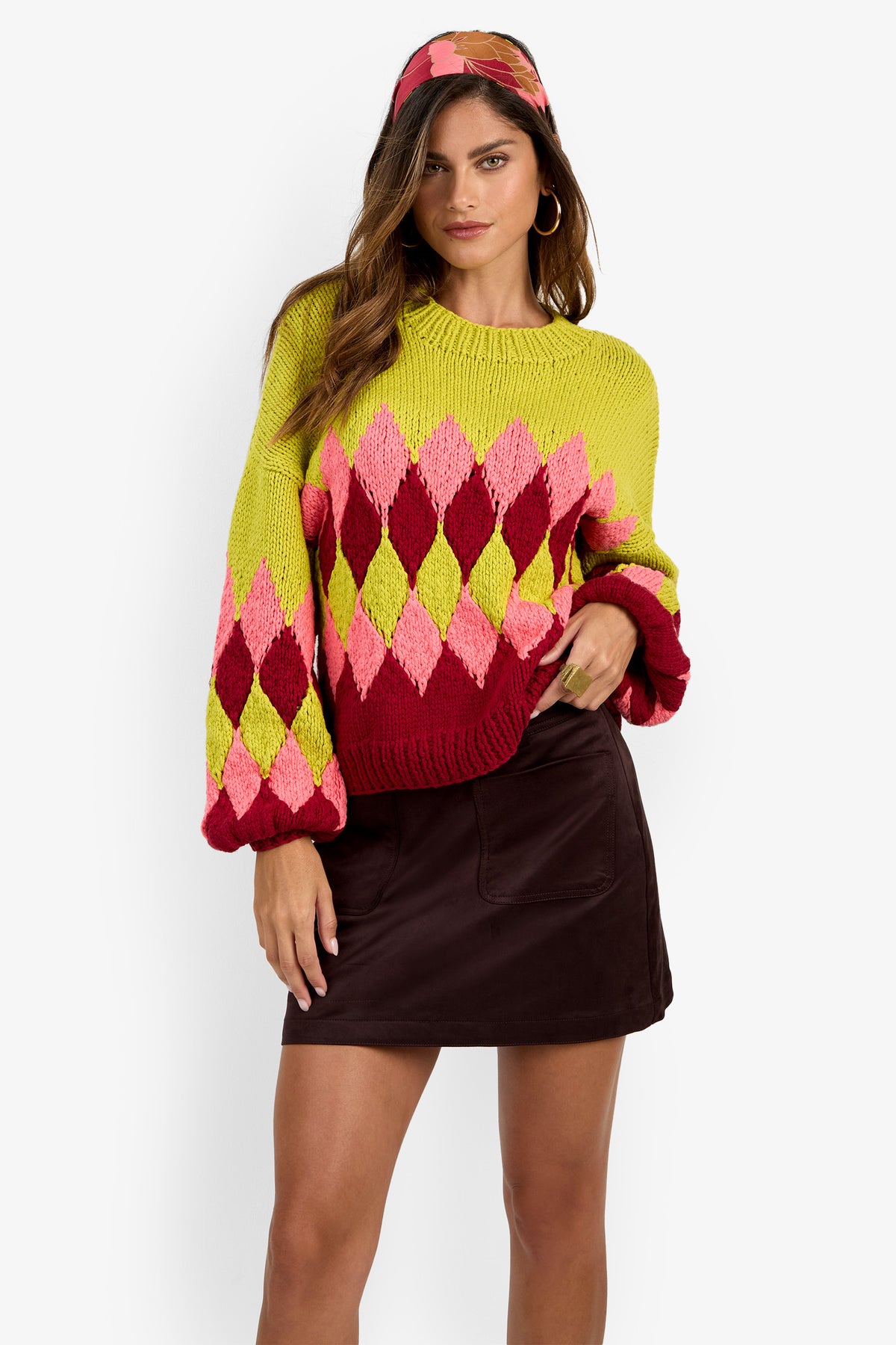 A woman poses with one hand on her hip against a white background, wearing the colorful Cavallari Sweater, complemented by a headband and a dark mini skirt.
