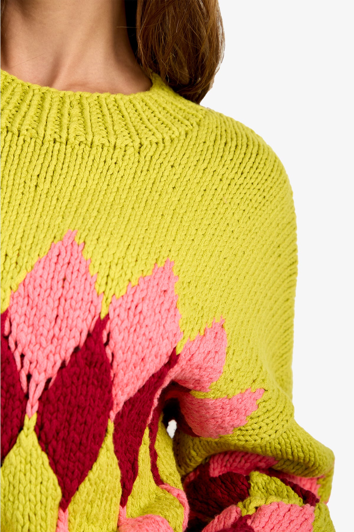 A close-up of a person wearing the Cavallari Sweater, a chunky knit yellow piece featuring a pink and red diamond pattern. Only part of their neck, shoulder, and hair are visible.
