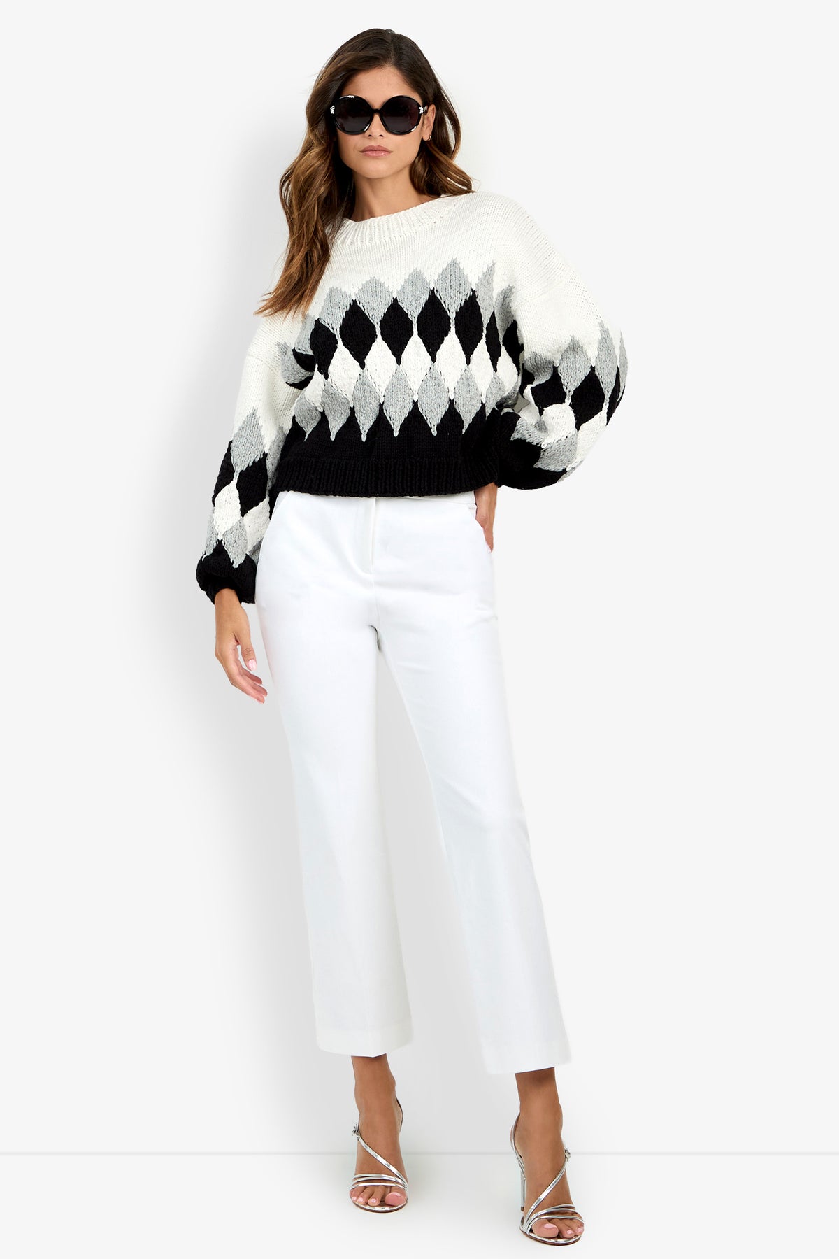 A woman wears a black, white, and grey argyle knit Cavallari Sweater with white trousers and silver heeled sandals.