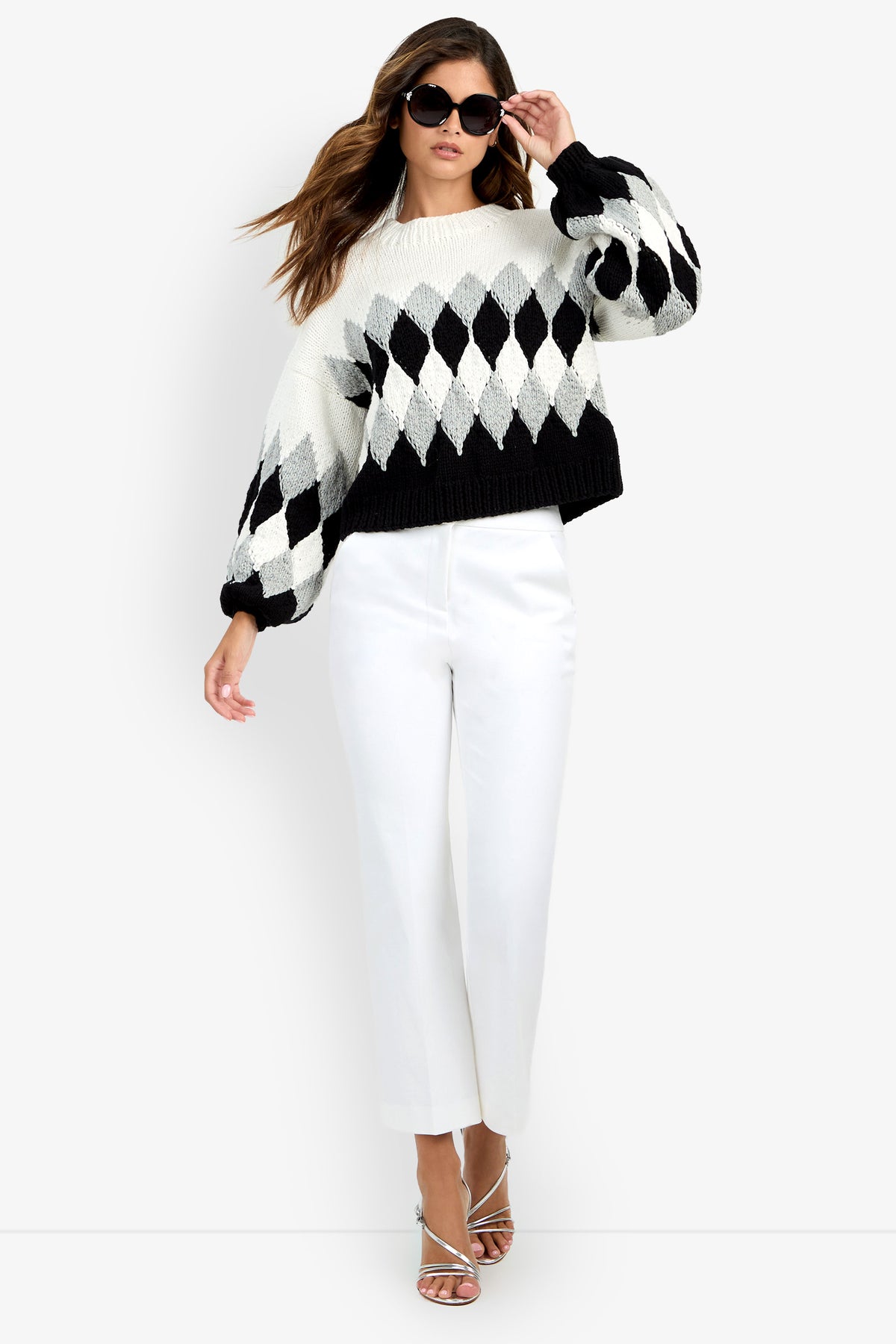 A person wearing a Cavallari Sweater featuring a black, white, and gray argyle pattern with long, puffed sleeves, paired with white trousers.