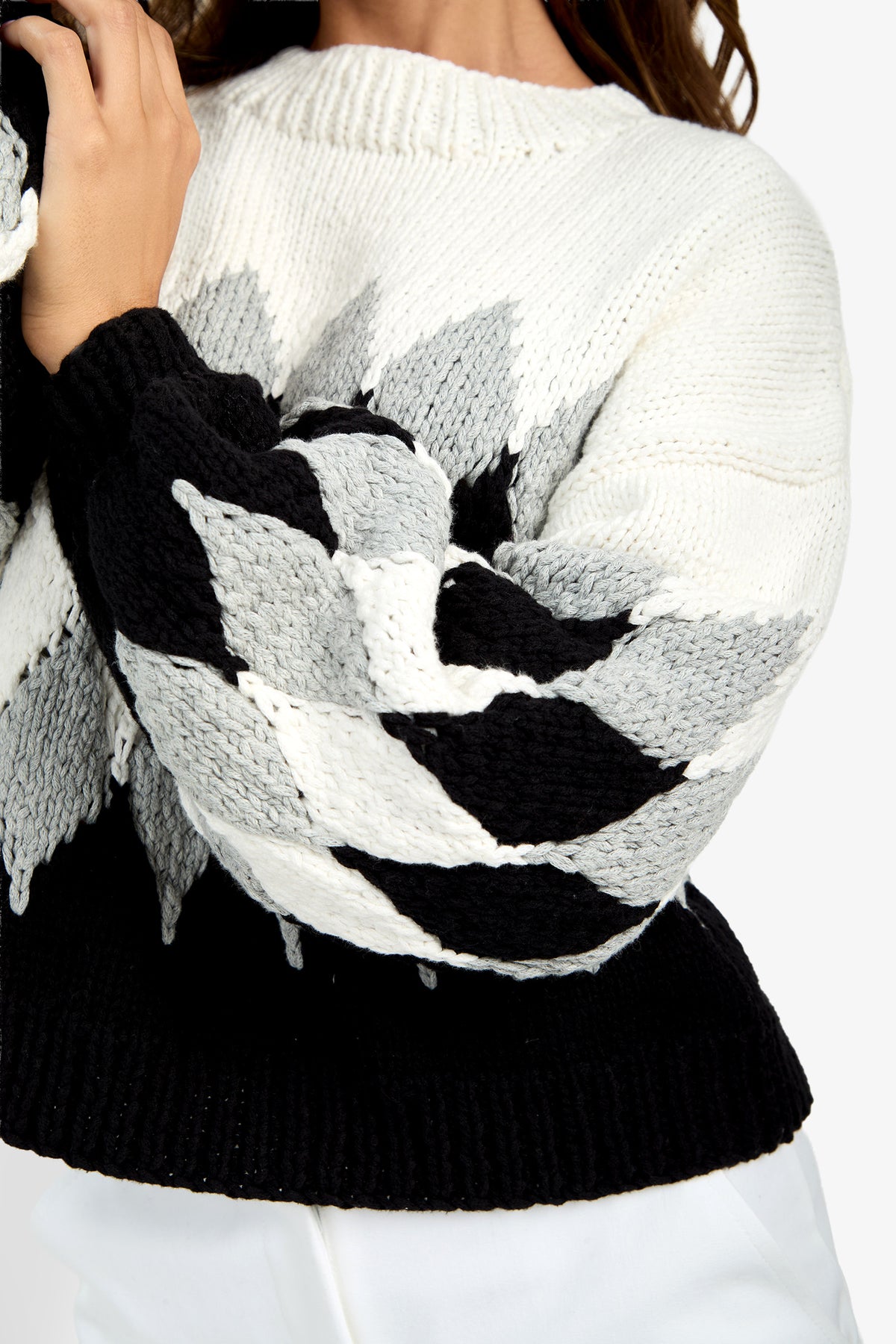 A person wears a Cavallari Sweater, a chunky knit style featuring a black, white, and gray geometric leaf-like pattern on the sleeves and a solid black hem.