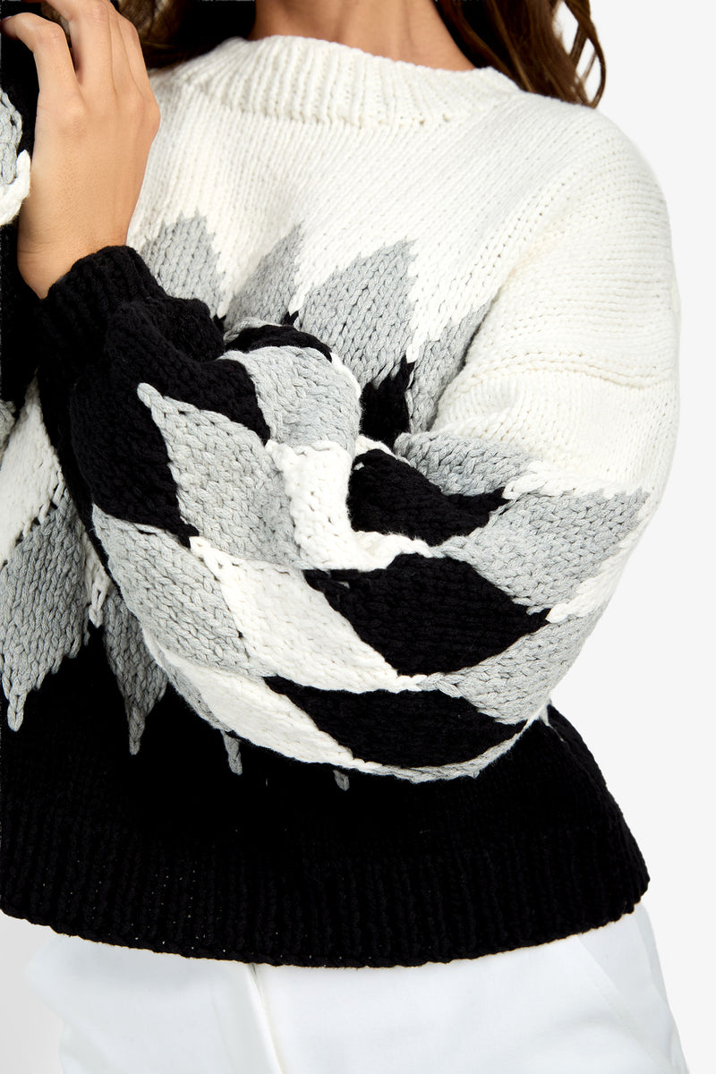 A person wears a Cavallari Sweater, a chunky knit style featuring a black, white, and gray geometric leaf-like pattern on the sleeves and a solid black hem.