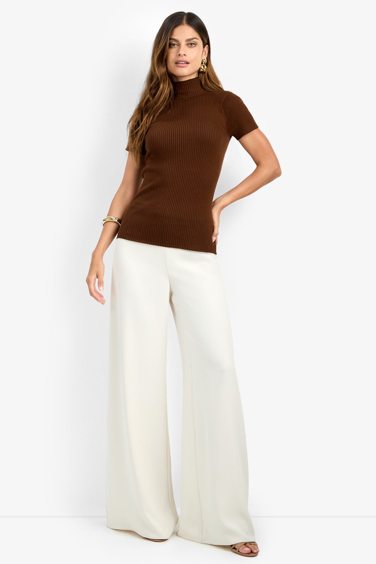 A woman stands against a plain white background, wearing the Armeni Mock Neck Top—a short-sleeve, ribbed brown turtleneck—paired with wide-leg white pants. She has long hair and accessorizes with gold earrings and bracelets.