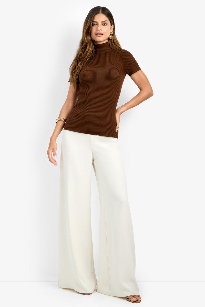 A woman stands against a plain white background, wearing the Armeni Mock Neck Top—a short-sleeve, ribbed brown turtleneck—paired with wide-leg white pants. She has long hair and accessorizes with gold earrings and bracelets.