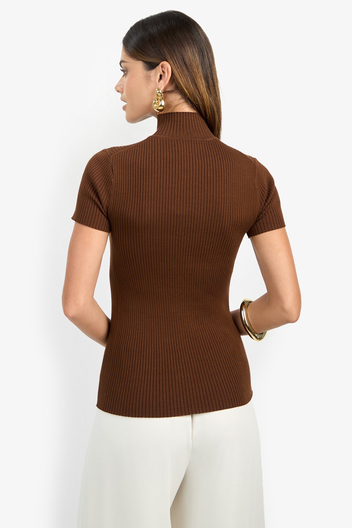 A woman is seen from behind wearing the Armeni Mock Neck Top in a ribbed brown fabric paired with white pants. She has straight brown hair, accessorized with large gold hoop earrings and gold bangles, set against a plain white background.