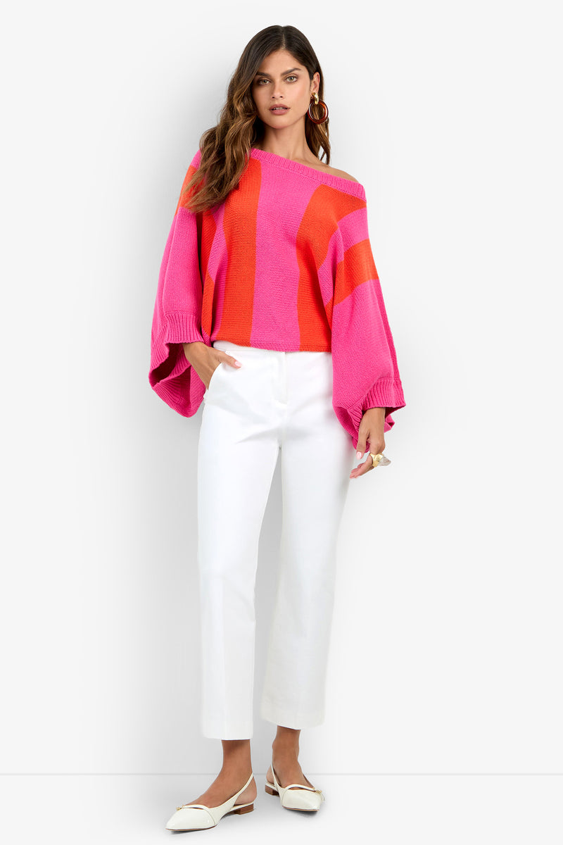 A woman wears an off-the-shoulder Horizon Sweater with wide pink and orange vertical stripes, paired with white trousers and white pointed-toe flats.
