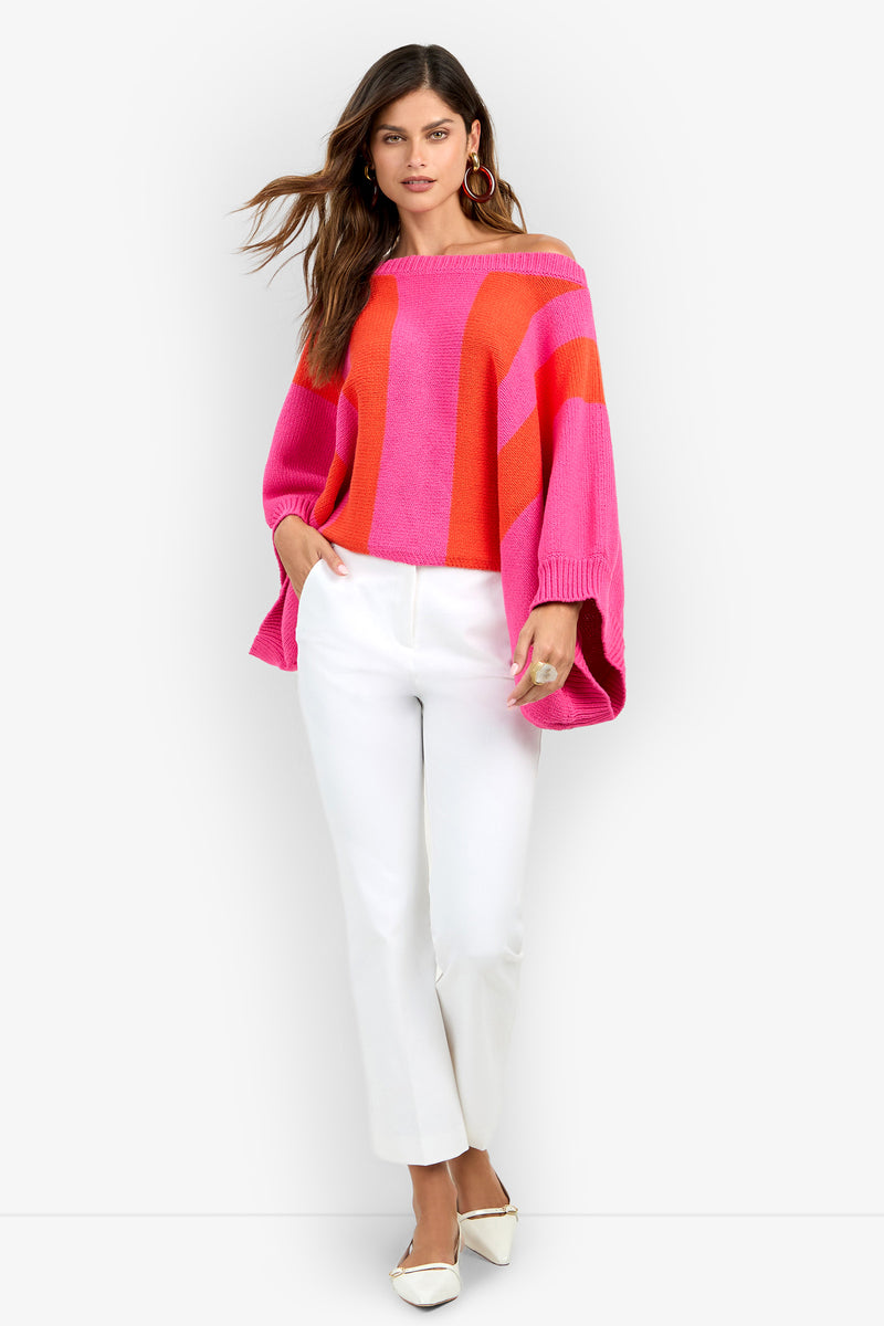A woman wears an off-the-shoulder Horizon Sweater with wide pink and orange vertical stripes, paired with white trousers and white flat shoes.