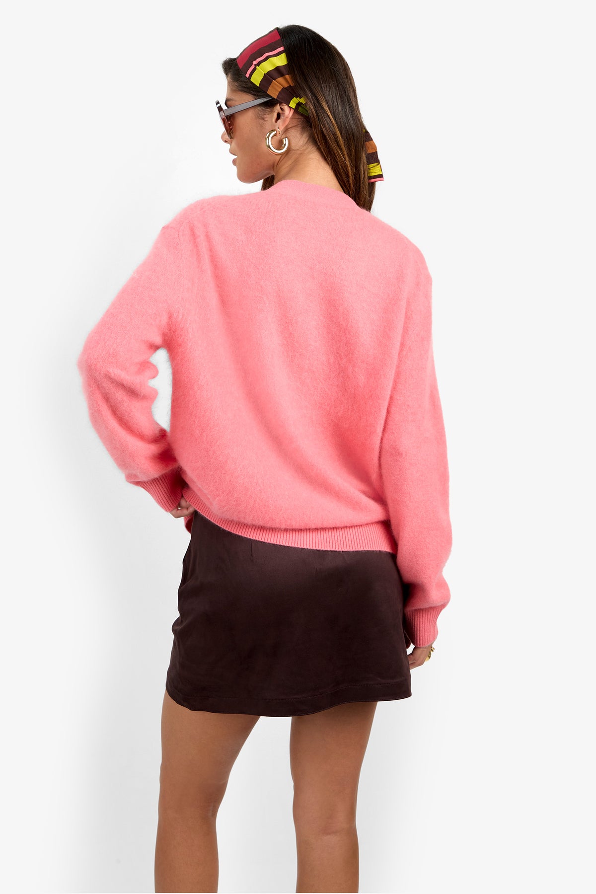 A woman stands against a plain white background, facing away while wearing the Milano Cardigan—a bright pink fuzzy sweater—paired with a dark brown mini skirt, sunglasses, gold hoop earrings, and a colorful striped scarf tied around her hair.