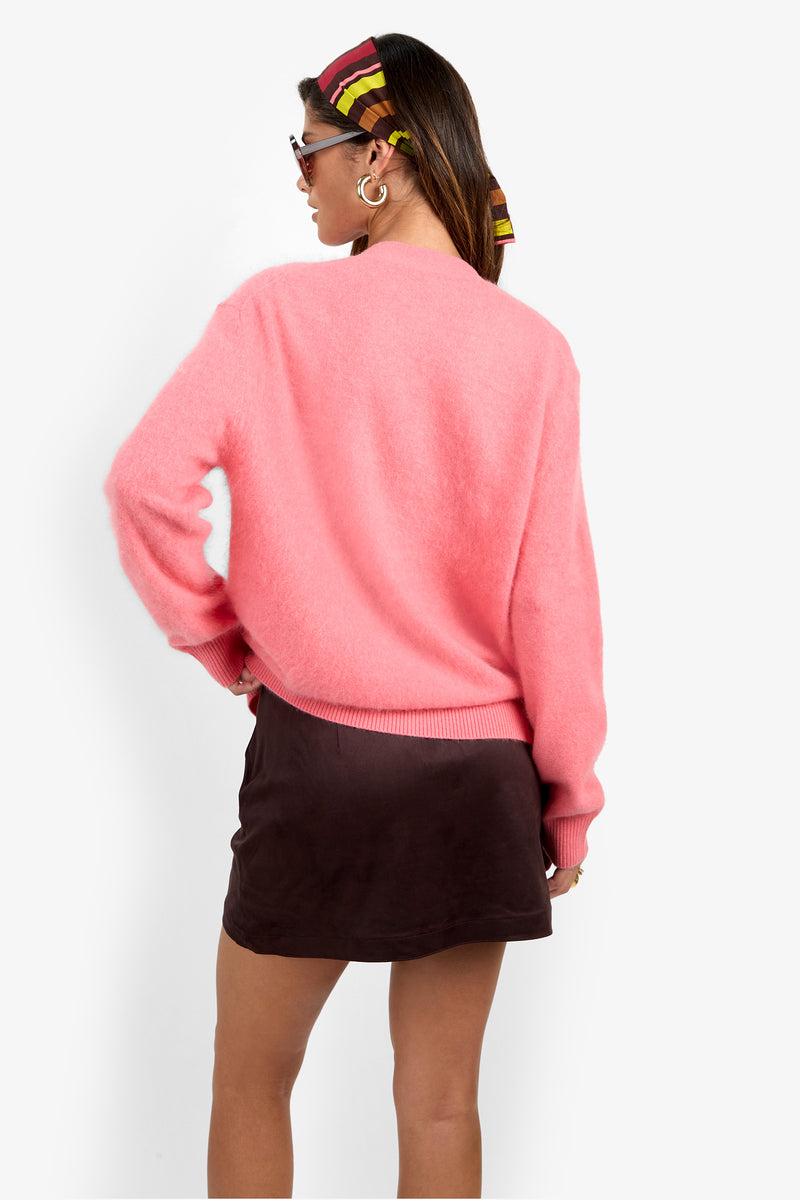 A woman stands against a plain white background, facing away while wearing the Milano Cardigan—a bright pink fuzzy sweater—paired with a dark brown mini skirt, sunglasses, gold hoop earrings, and a colorful striped scarf tied around her hair.
