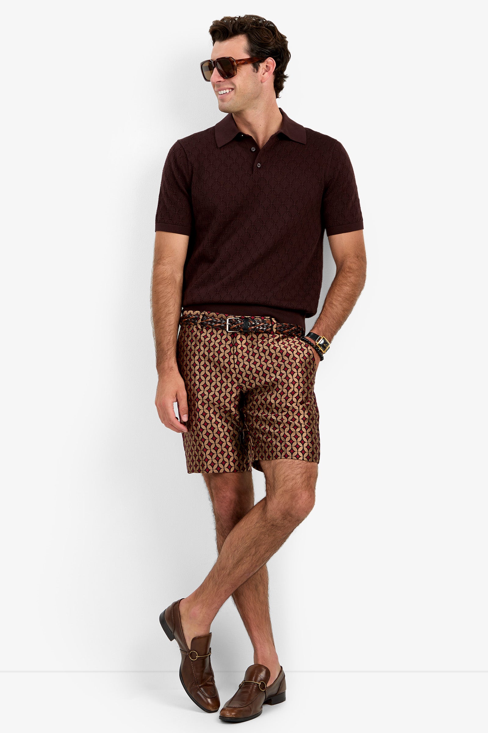 A man wearing sunglasses, the Waylen Polo in short-sleeve maroon, patterned shorts, a brown belt, a watch, and brown loafers stands against a plain white background, looking to the side and smiling.