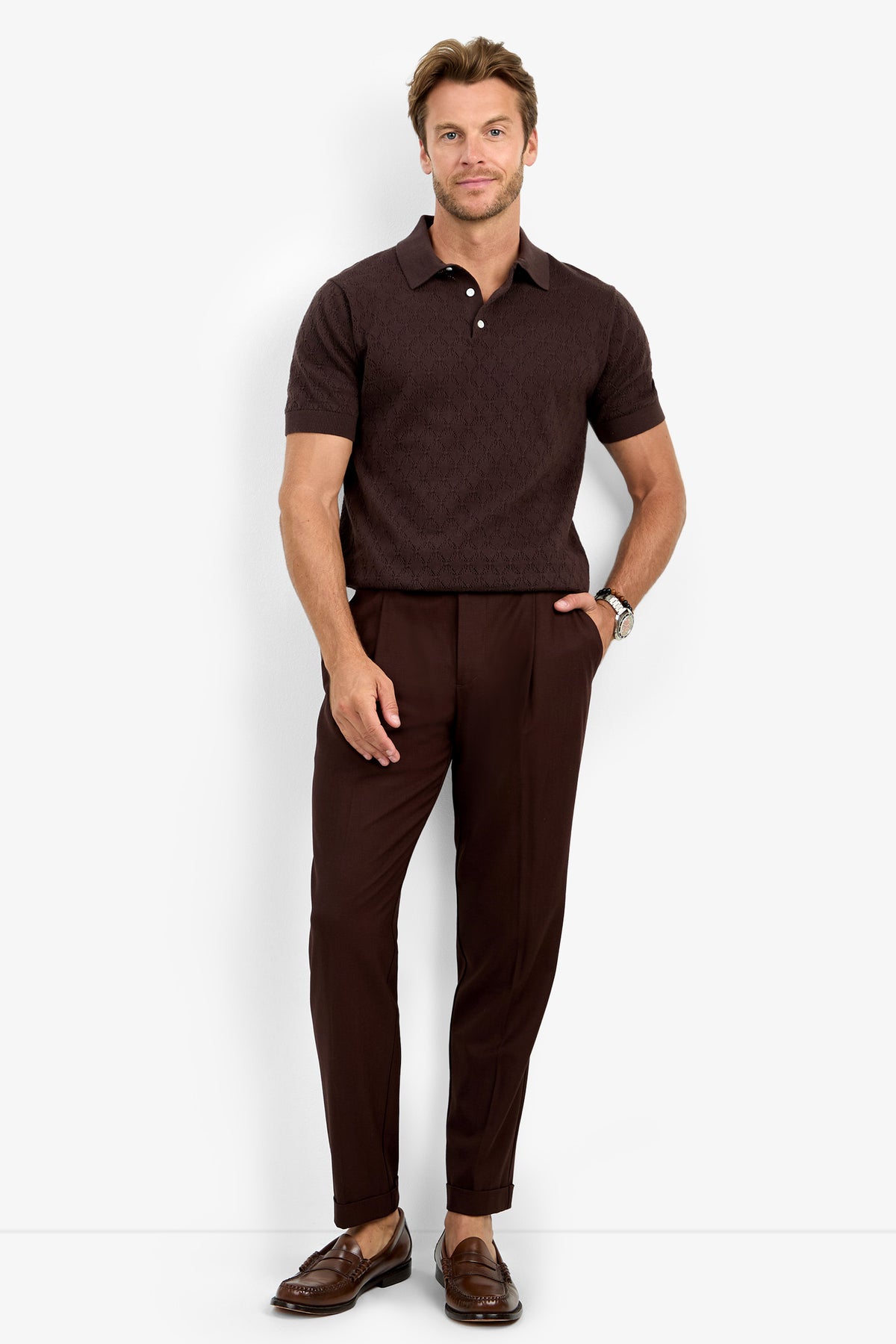 A man stands against a white background wearing the WAYLEN SS POLO, paired with brown pleated pants and brown loafers. He has one hand in his pocket and is smiling slightly.