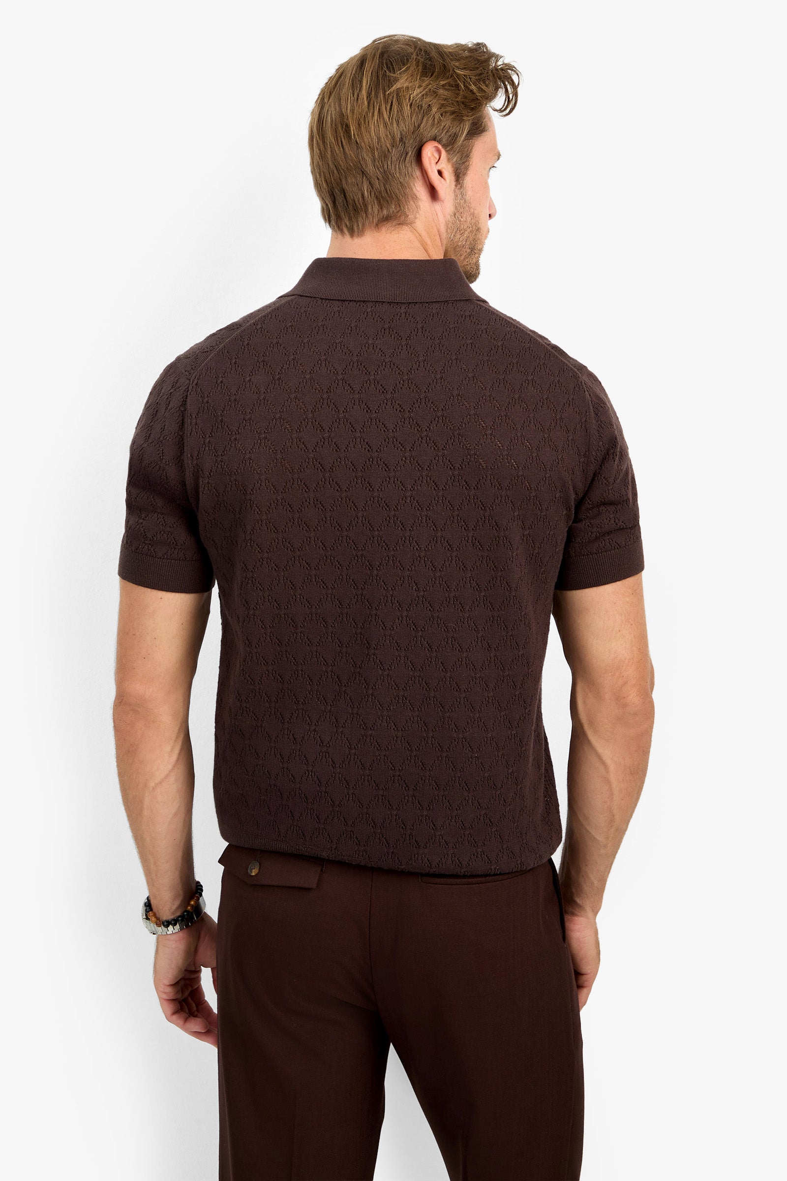 A man with light brown hair is seen from the back, standing against a plain white background, wearing the WAYLEN SS POLO— a short-sleeve, textured dark brown polo shirt—paired with matching brown pants.