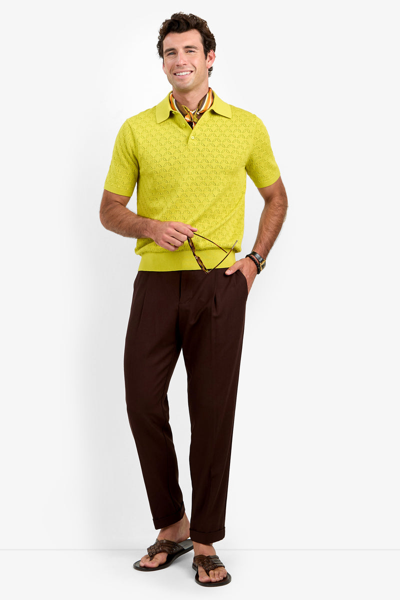 A smiling man stands against a plain white background, exuding relaxed style in the Waylen Polo—a yellow short-sleeve sweater—paired with brown pants, sandals, and a patterned scarf around his neck. He holds sunglasses in one hand, completing the effortlessly stylish look.