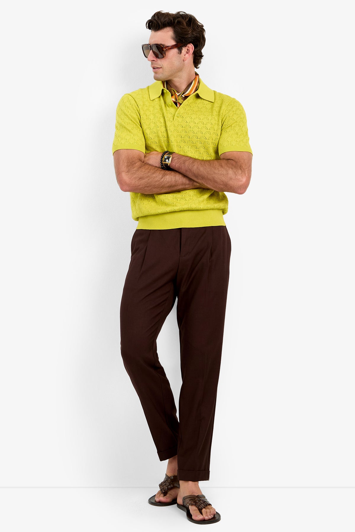A man wearing the Waylen Polo, sunglasses, brown pants, a watch, and flip-flops stands with arms crossed against a white background.