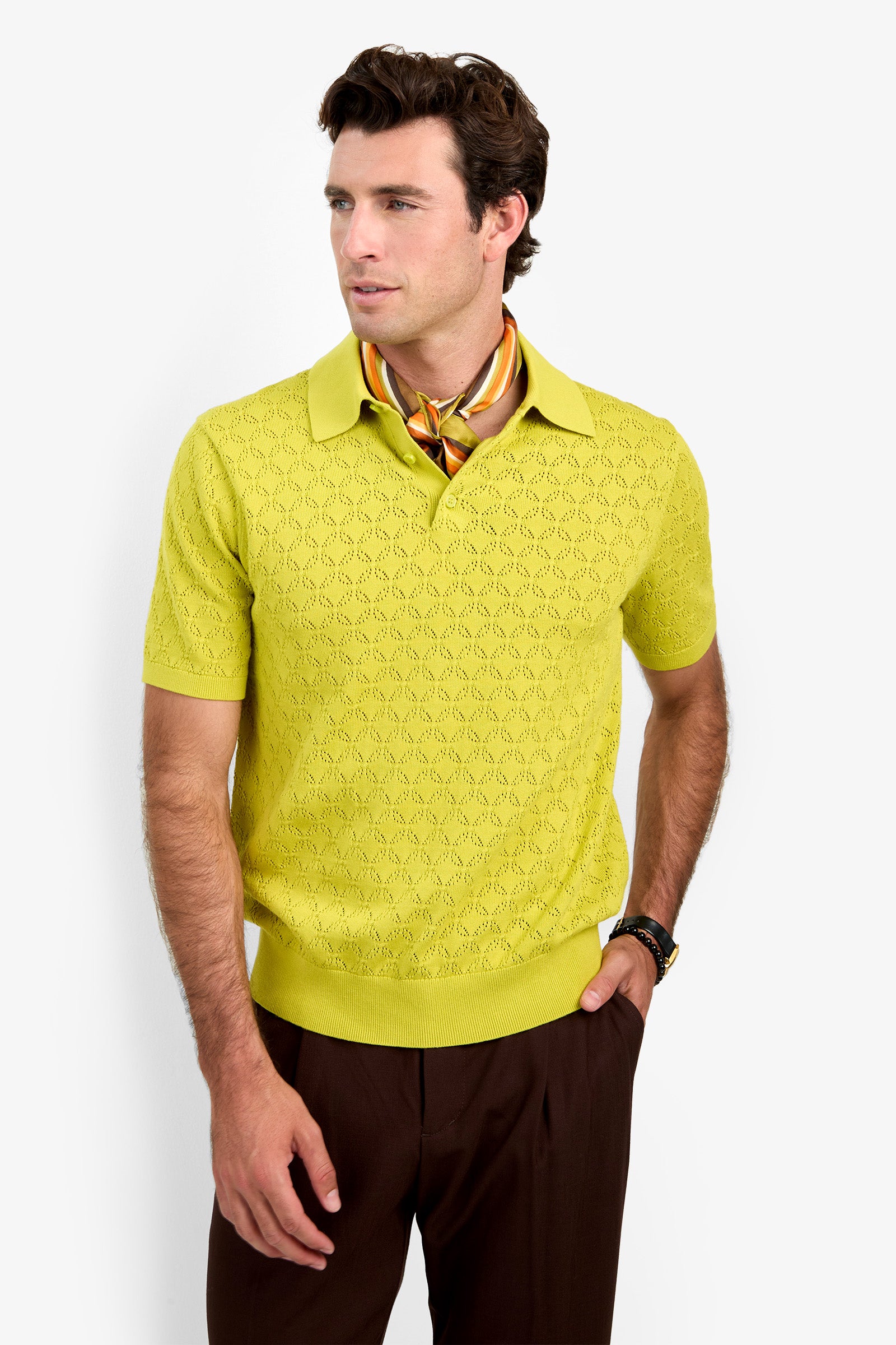 A man wearing the yellow Waylen Polo, brown pants, a patterned silk scarf, and a wristwatch stands against a white background, looking to the side with one hand in his pocket.