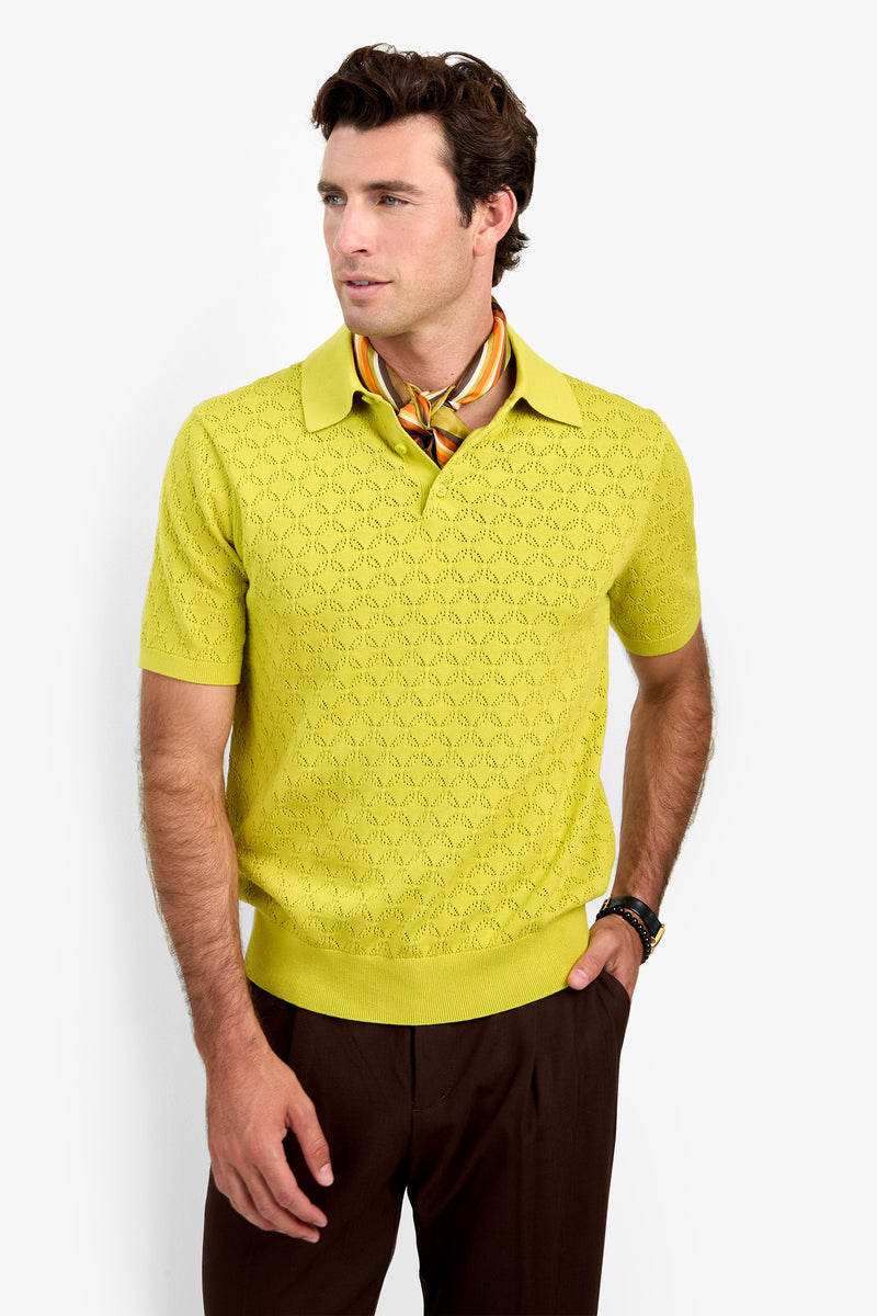A man wearing the yellow Waylen Polo, brown pants, a patterned silk scarf, and a wristwatch stands against a white background, looking to the side with one hand in his pocket.