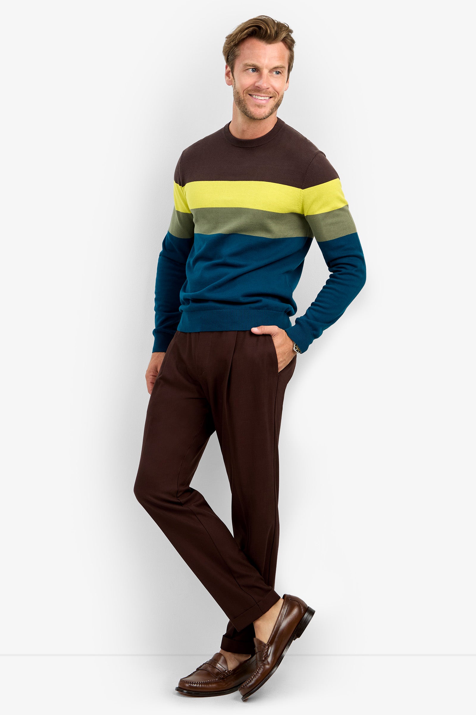 A man stands and smiles, wearing the ROEMER sweater—a color-block design crafted from 100% cotton with bold brown, yellow-green, and blue stripes. He pairs it with brown pants and dress shoes, posing casually with one hand in his pocket against a simple backdrop.