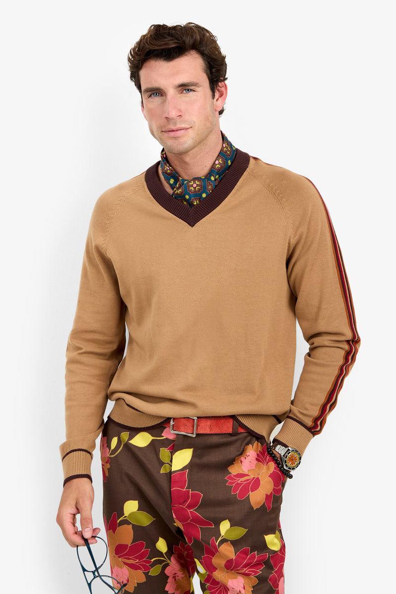 A man wearing the Harvey Sweater—a tan sweater with maroon trim—paired with a patterned shirt and brown pants featuring red and yellow floral prints, stands against a white background holding eyeglasses and wearing a watch.