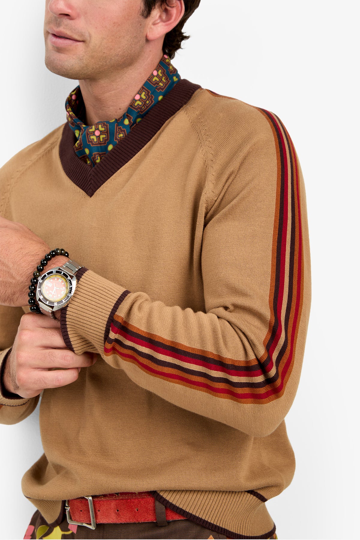 A man wearing the Harvey Sweater, featuring tan fabric with red and brown stripes on the sleeves. He pairs it with a patterned shirt underneath and accessorizes with a watch and beaded bracelets. His face is partially visible as he stands with his arms crossed.