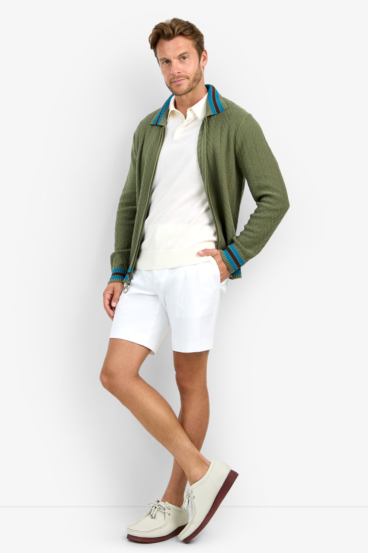 A man poses against a white background wearing the ROBERTO ZIP CARDIGAN in olive green, paired with a white polo shirt, white shorts, and light-colored shoes with brown soles.