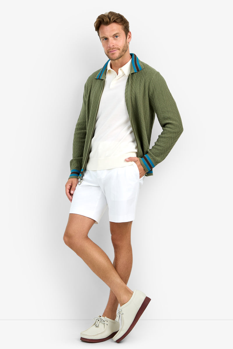 A man poses against a white background wearing the ROBERTO ZIP CARDIGAN in olive green, paired with a white polo shirt, white shorts, and light-colored shoes with brown soles.