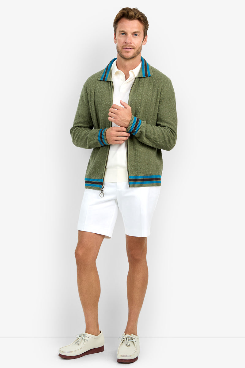 A man stands facing forward against a plain light background, wearing the ROBERTO ZIP CARDIGAN—a light green cardigan with blue stripes—paired with a white polo shirt, white shorts, and off-white shoes; he holds his jacket with one hand.