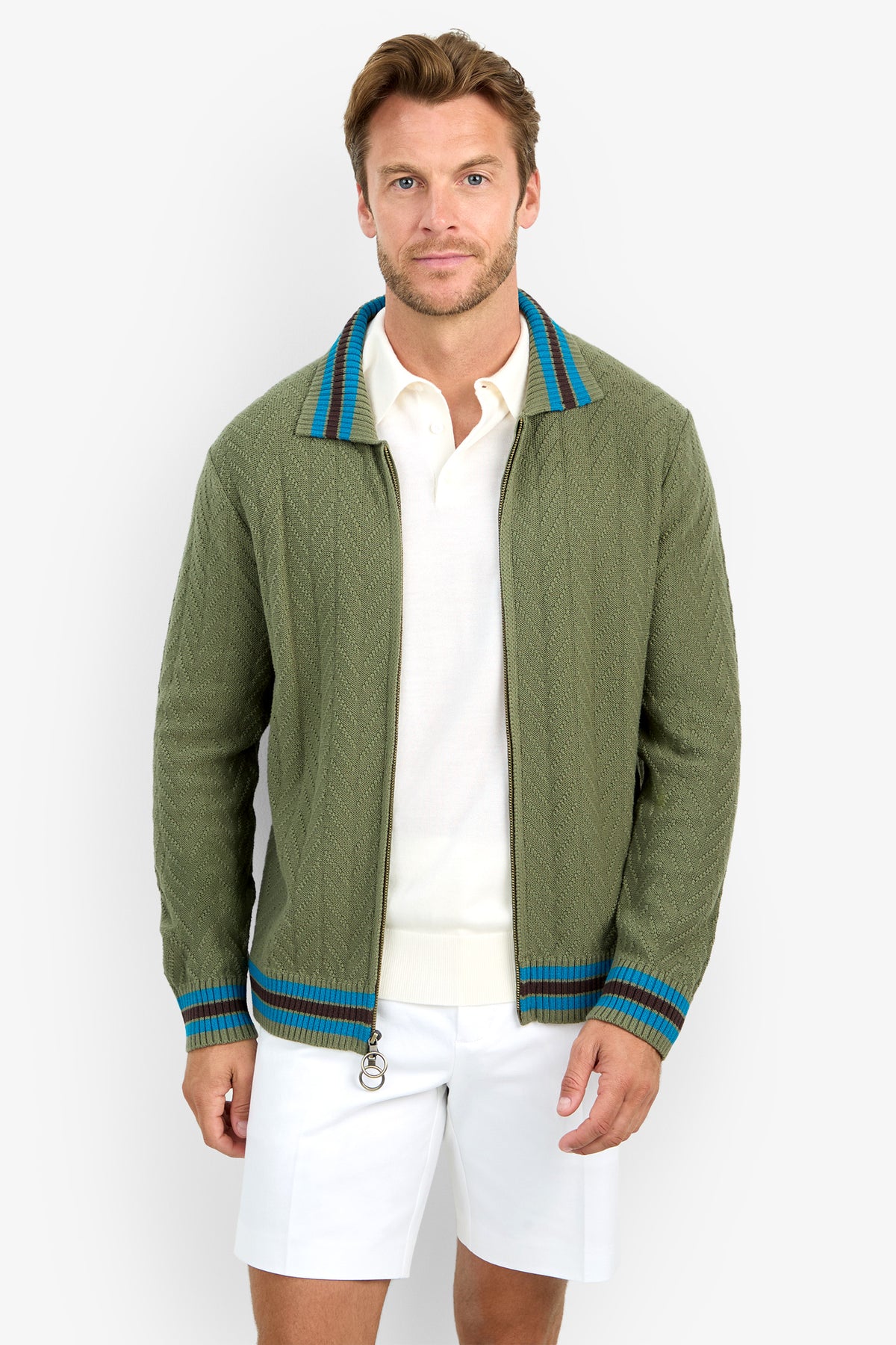 A man wearing the ROBERTO ZIP CARDIGAN, featuring a green knit with blue and brown striped collar and cuffs, paired with a white collared shirt and white shorts, stands facing forward against a plain white background.