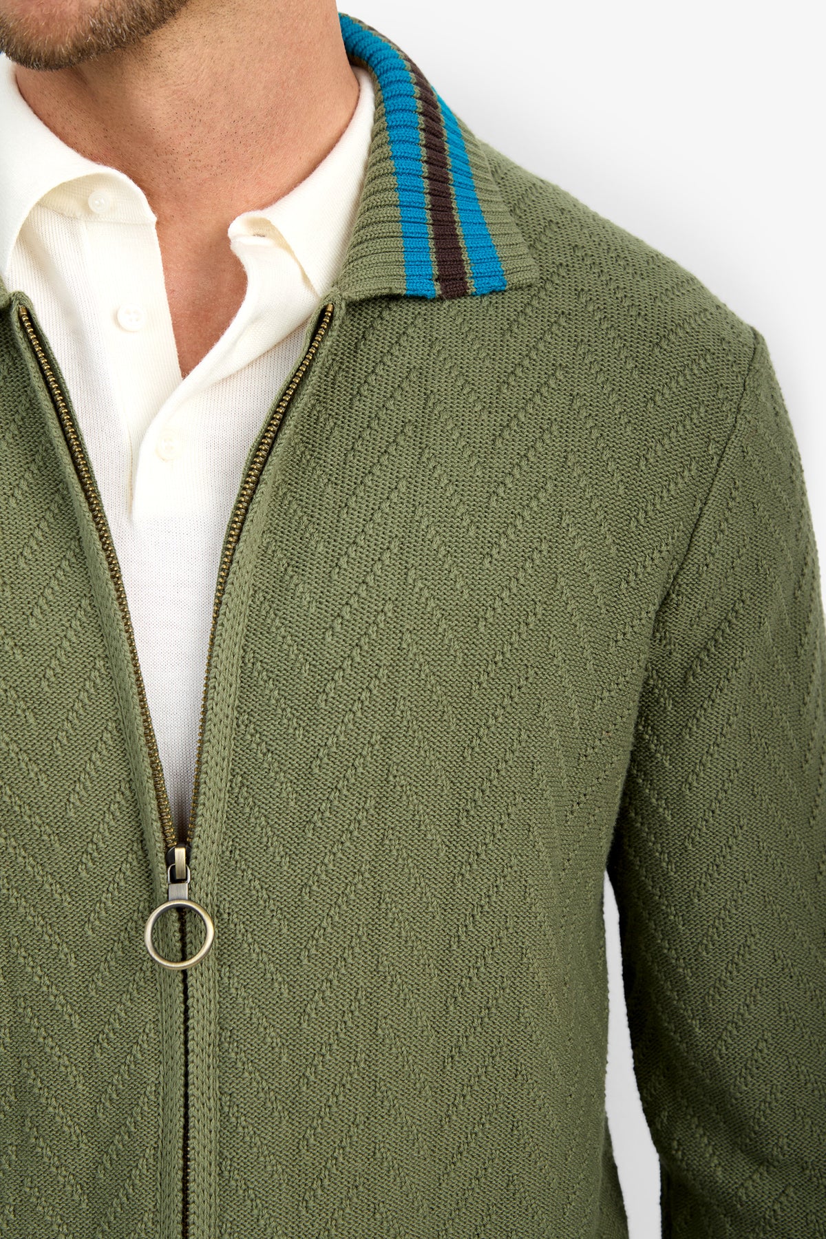 A close-up of a person wearing the ROBERTO ZIP CARDIGAN, an olive green textured zip-up jacket with a blue and brown striped collar, layered over a white shirt. Only the lower face, neck, and upper torso are visible.