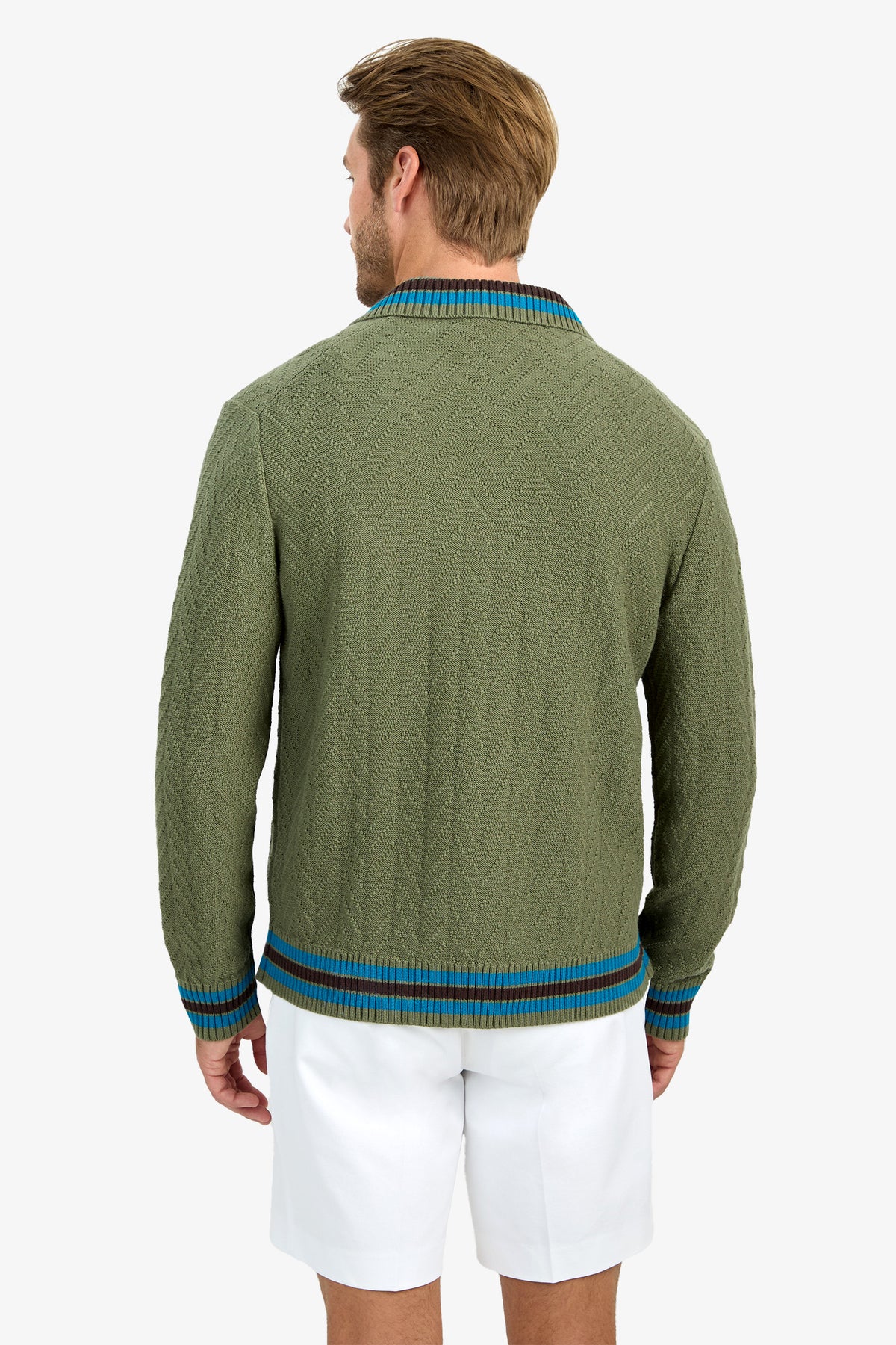 A man with light brown hair, seen from behind, wears the ROBERTO ZIP CARDIGAN—a textured olive green sweater with blue and brown trim—paired with white shorts.