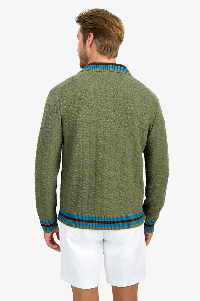 A man with light brown hair, seen from behind, wears the ROBERTO ZIP CARDIGAN—a textured olive green sweater with blue and brown trim—paired with white shorts.