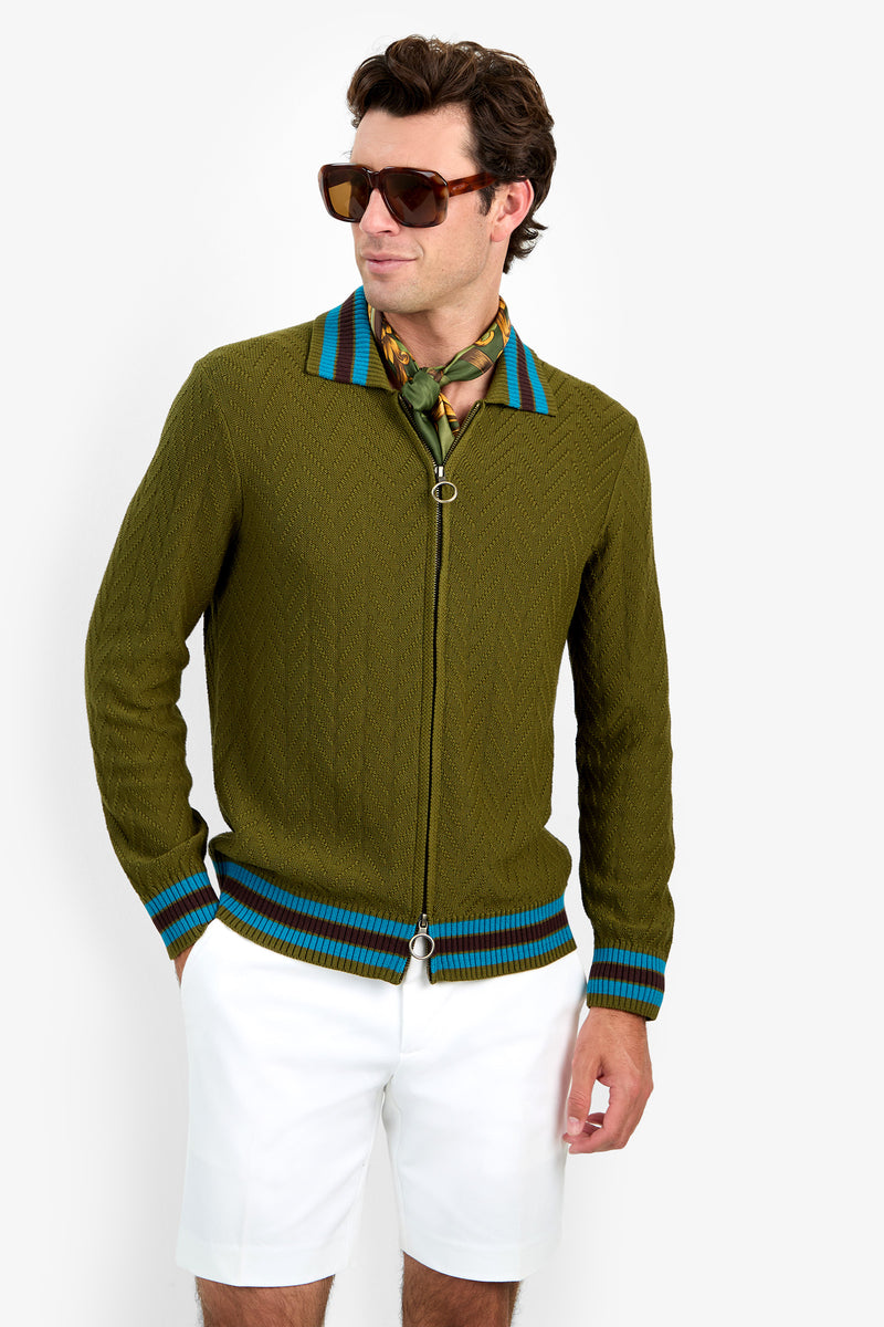 A man in sunglasses wears the Roberto Zip Cardigan, an olive green zip-up sweater featuring blue stripes on the collar and cuffs. He pairs it with a patterned scarf and white shorts while standing against a plain white background.