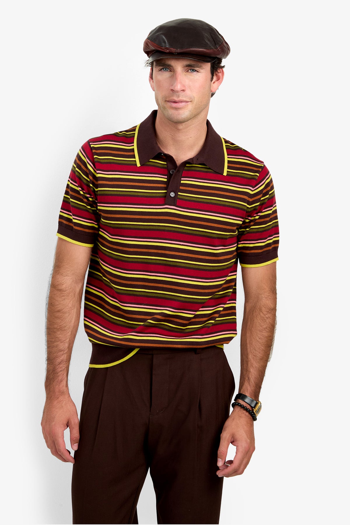 A man wearing a brown flat cap, the Bayard Polo—a short-sleeve striped shirt in brown, red, and yellow—and dark brown pants stands against a plain light background.