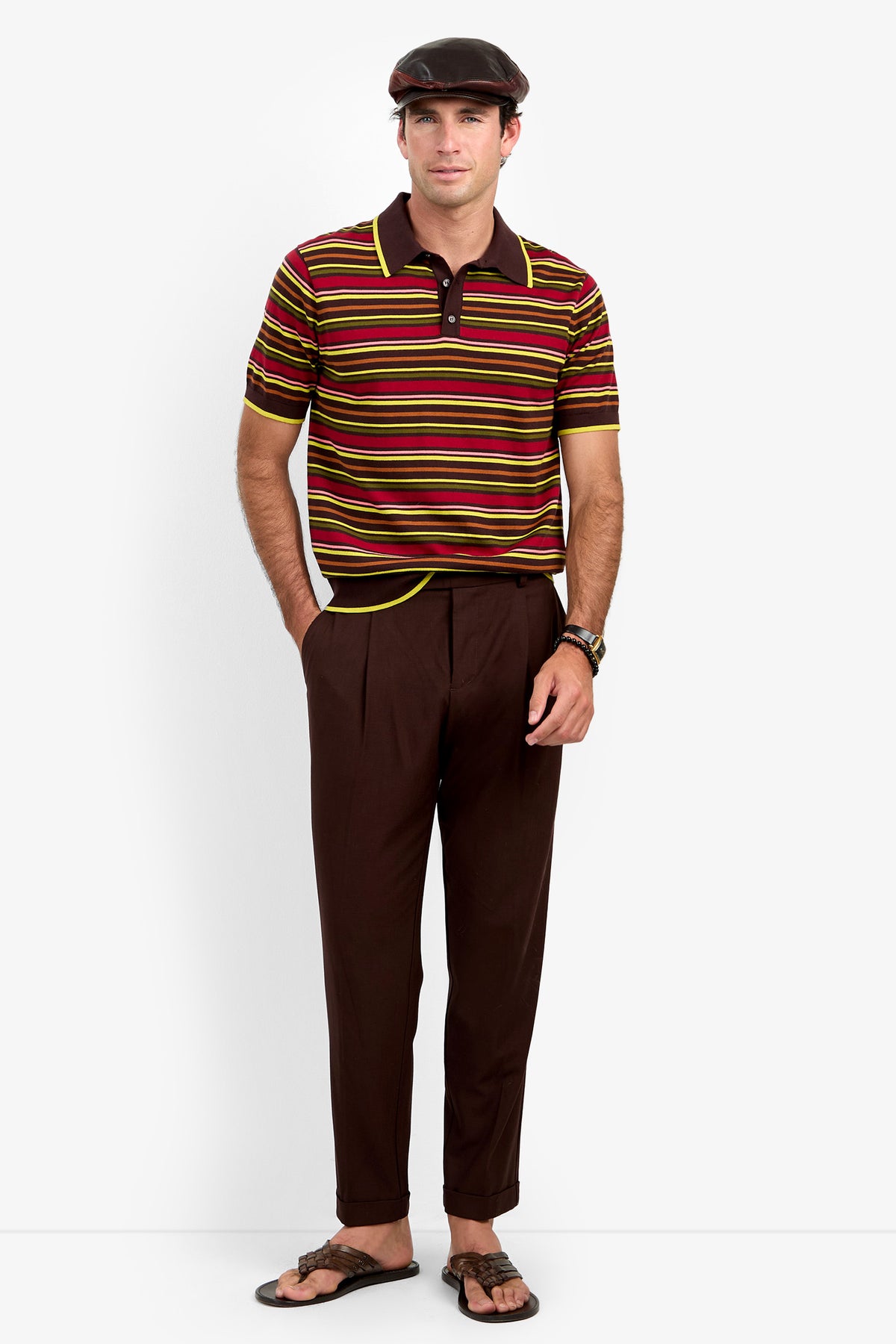 A man wearing the Bayard Polo, brown trousers, a dark flat cap, and sandals stands with one hand in his pocket against a plain white background.