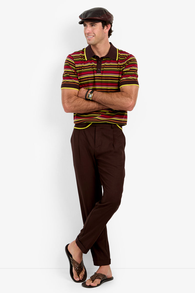A man wearing the Bayard Polo, brown trousers, a dark brown cap, and sandals stands with his arms crossed and smiles.
