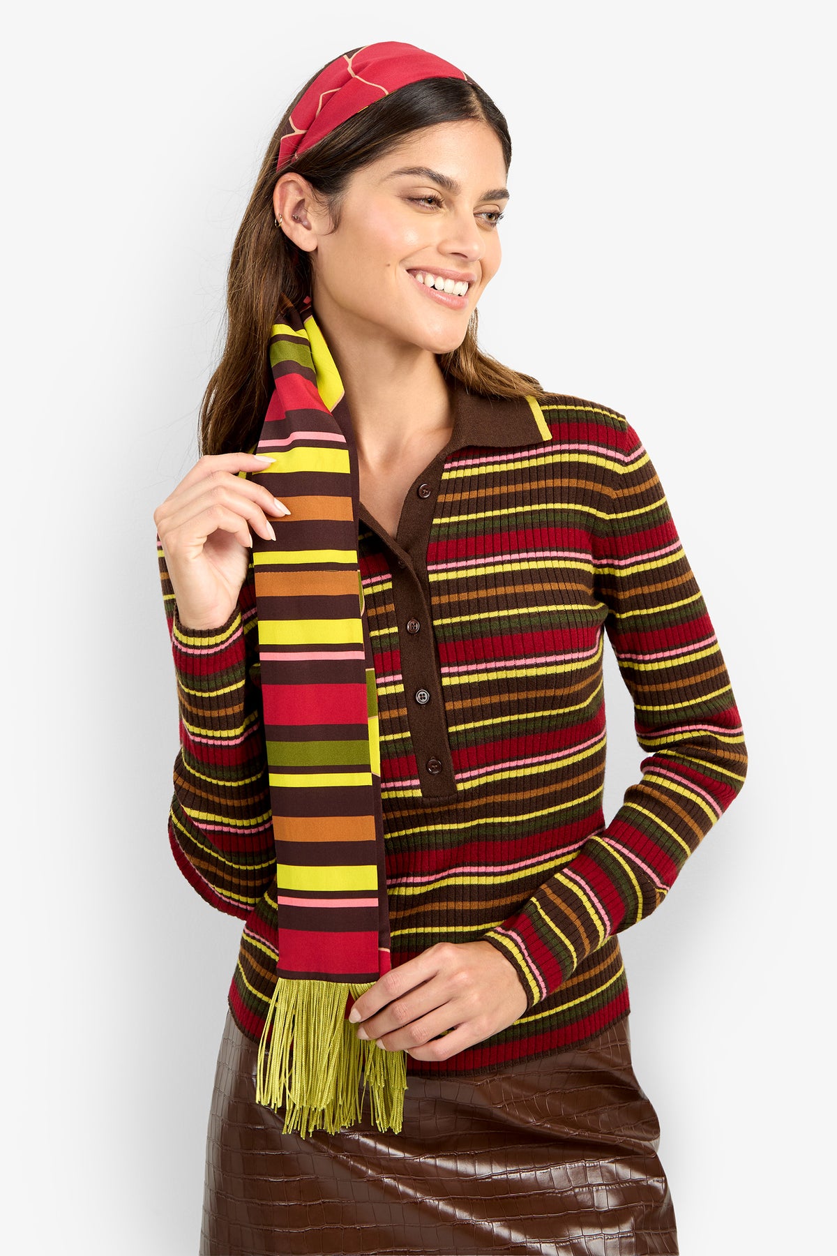 A woman with long brown hair smiles and looks to the side as she poses against a plain white background, wearing a brown skirt and a colorful striped sweater paired with the SERA FRINGE SCARF.
