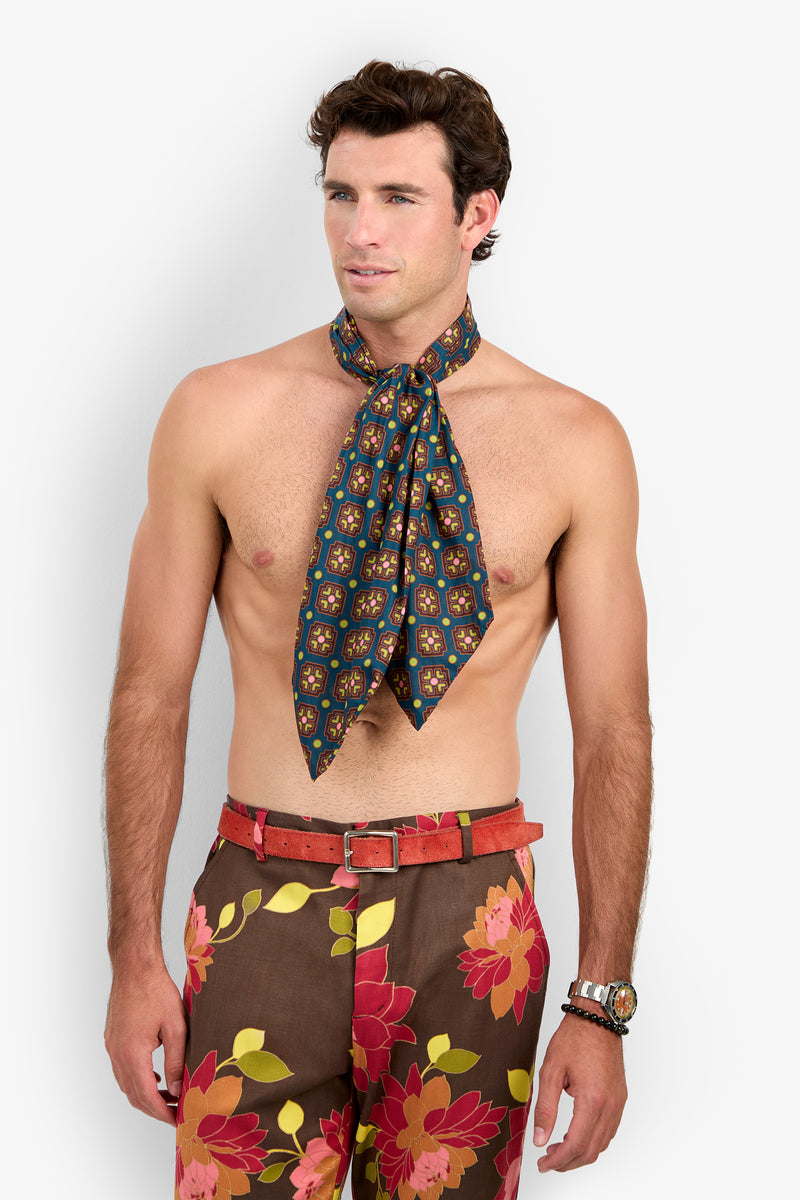 A shirtless man wearing floral pants with a red belt and an Ascot patterned neck scarf stands against a plain white background, looking slightly to the side.